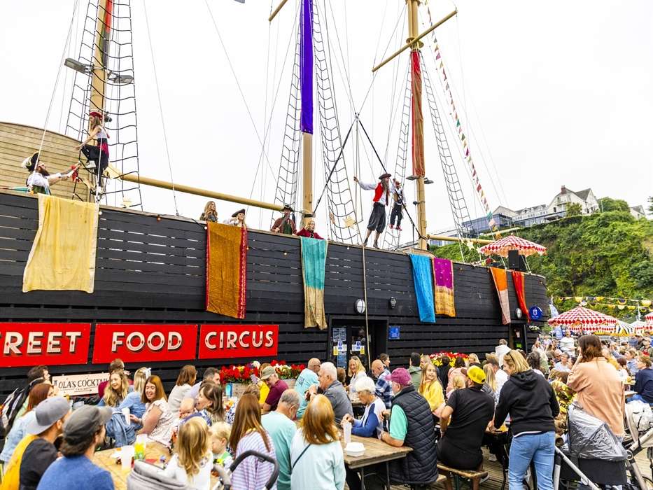 Crowds of people dining at long tables next to a large black vintage pirate ship with Street Food Circus banners at Saundersfoot Harbour