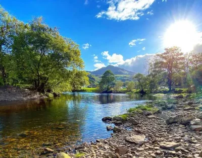 The river Llugwy running over stones below wooded banks and a mountain at Dolgam Campsite
